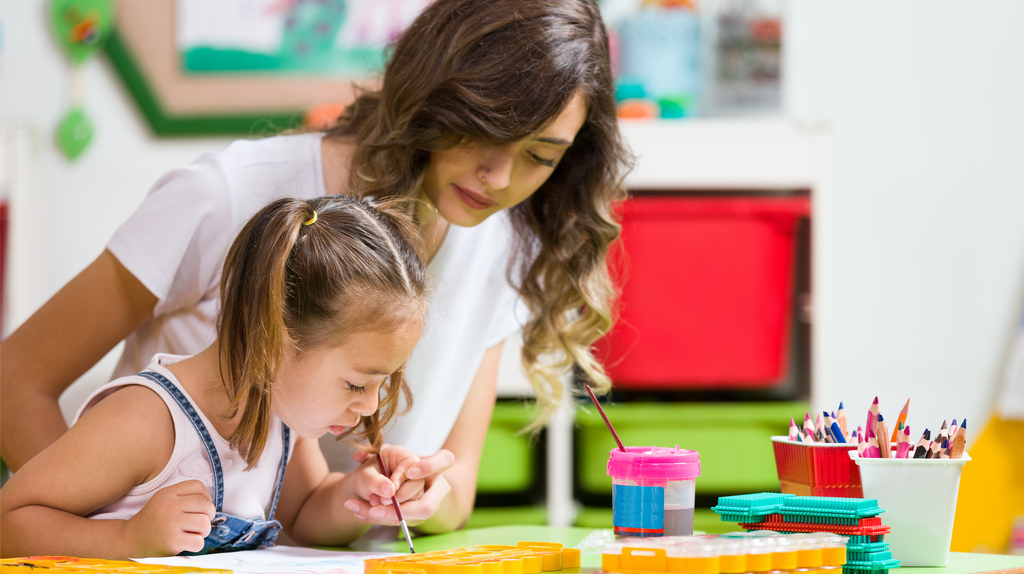 A teacher assisting a child to paint in a nursery.